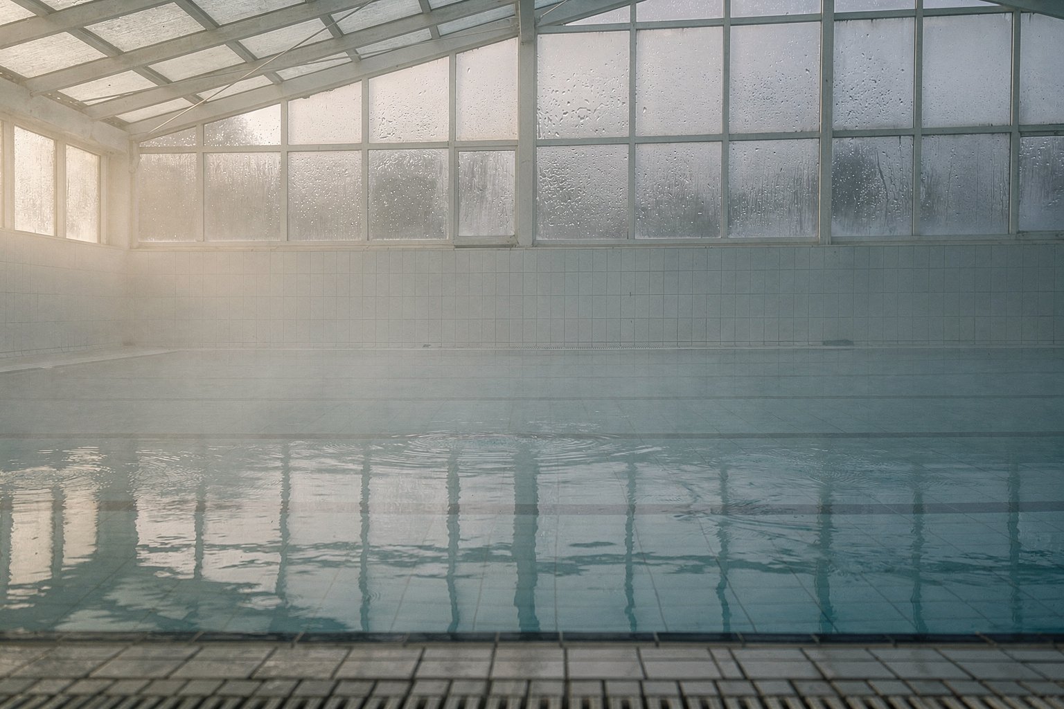 Un bassin de piscine communale désert au petit matin, la buée sur la verrière et l’eau immobile.